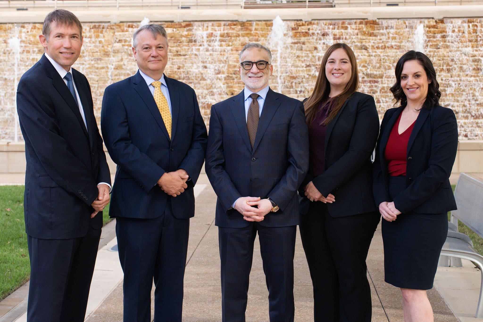 The five attorneys of The Mussalli Law Firm standing together in navy suits in front of a stone fountain wall